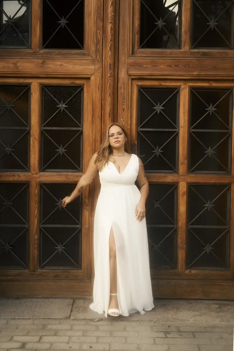 Gisele sozinha, de vestido branco, diante de um imenso portão de madeira trabalhada
