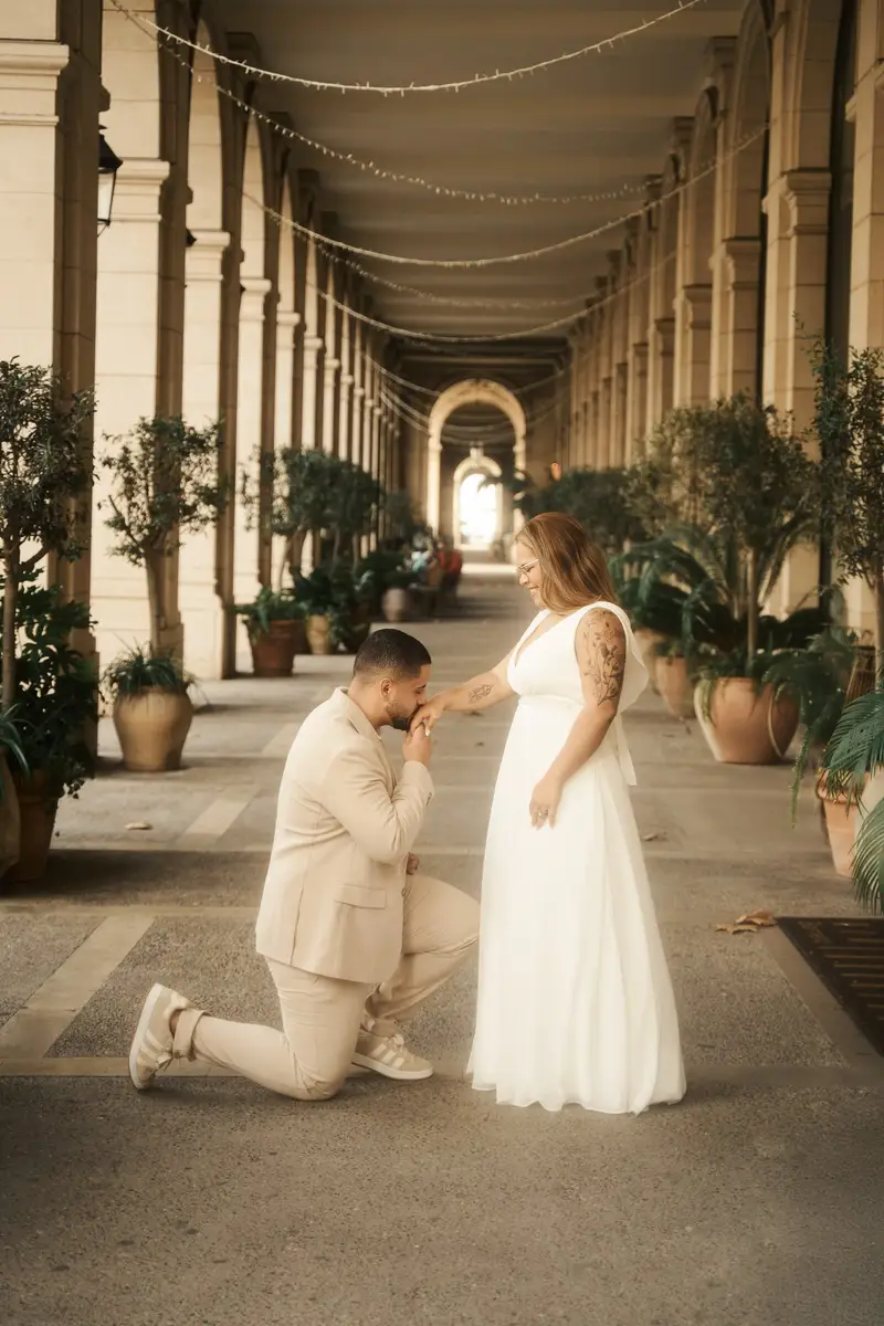 Victor ajoelhado beijando a mão de Gisele, numa galeria de colunas com vasos de plantas